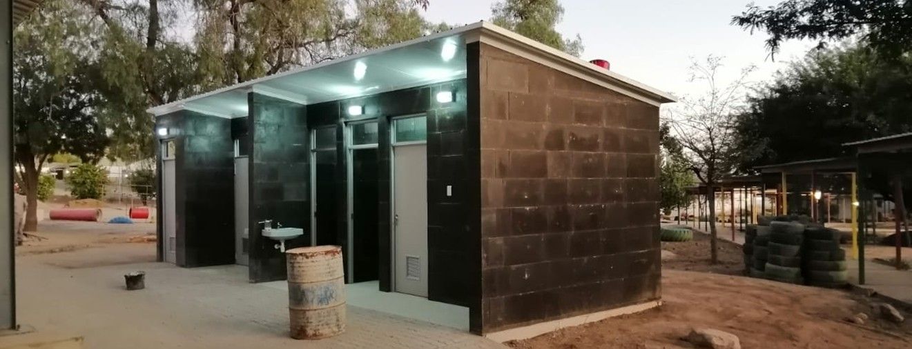 Die neue Sanitäranlage der Dagbreek School aus dunklen Polyblocks im Schatten von Bäumen