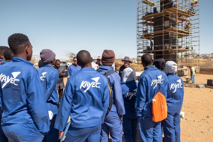 Gruppe von Lernenden in blauer Arbeitskleidung mit KAYEC-Aufdruck auf dem Gelände, im Hintergrund der eingerüstete Turm