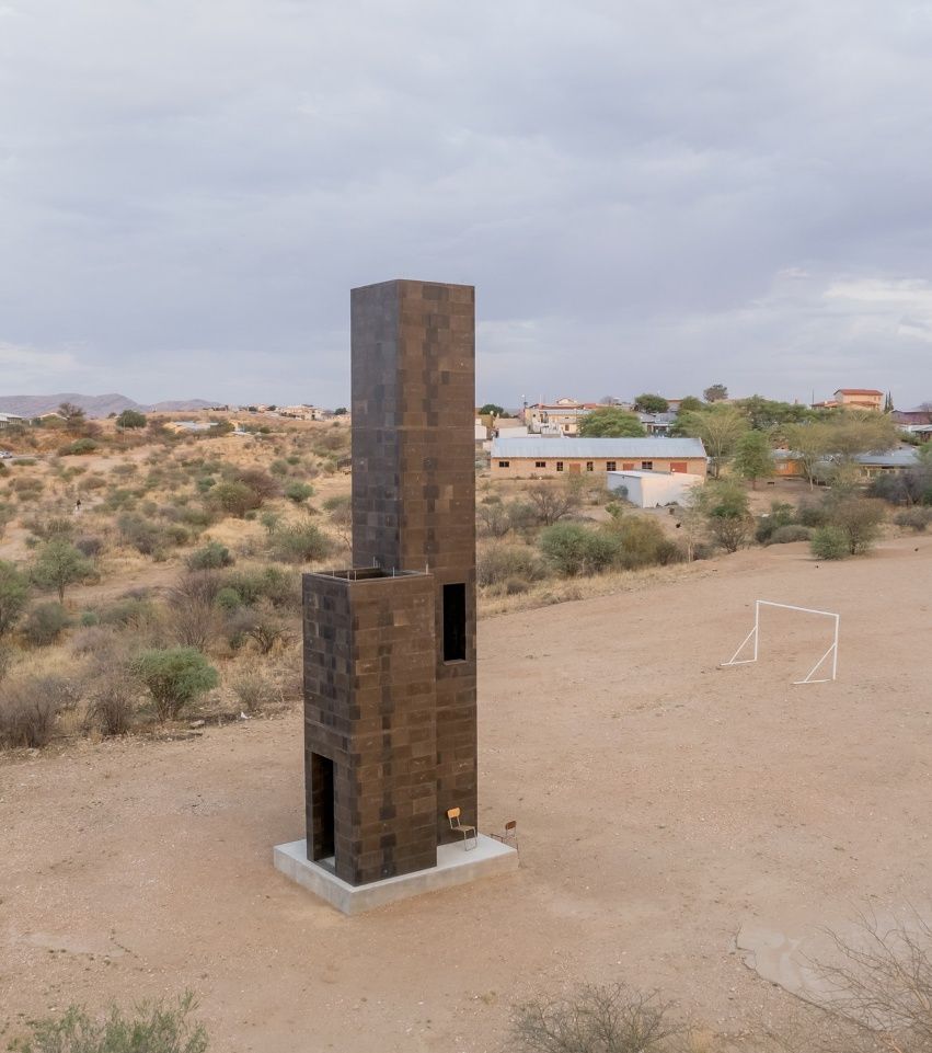 Der Material-Test-Turm aus dunklen Steinen auf sandigem Gelände, im Hintergrund ein Fußballtor
